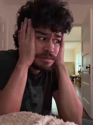 A man, appearing distressed, clutches his head with both hands, his eyes wide with an expression of concern or sorrow. His curly hair and facial hair add to the intensity of his expression. The background is a blurred interior with implied natural light.