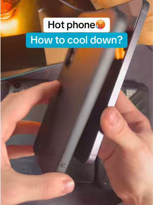 A person's hands are holding a smartphone at an angle, showcasing the back of the phone. Overlay text in a speech bubble, top-center, reads, 'Hot phone', and the secondary text, in a rectangle overlay at the bottom-center, reads, 'How to cool down?' The background is blurred, showcasing a wooden table.