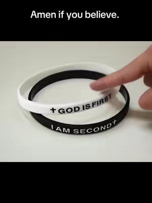 The image features a close-up shot of two silicone wristbands, one white and one black, placed on a light-colored surface. The white wristband is positioned atop the black one, with a finger gently touching it. The white wristband has the text 'GOD IS FIRST' and a cross symbol printed on it. The black wristband has the text 'I AM SECOND' printed on it. The text 'Amen if you believe.' is present in a black bar at the top center of the image with black background.