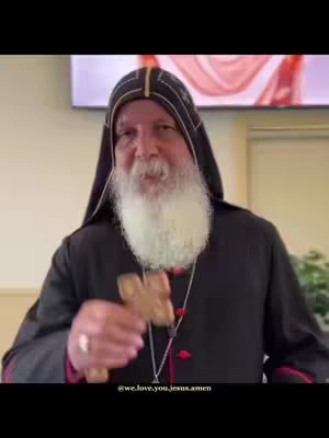 The image shows a man with a long white beard and a black and gold hat, holding a wooden cross. He is wearing a black robe. The background is blurred, but appears to be a room with a screen and green plants.
