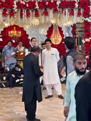 The image shows a gathering in a decorated hall. Prominently positioned just left of the image's center stands a man in white traditional attire, his arms at his sides. To his left is a man in a black suit. Behind them, a group of other men are visible; some appearing to be singing or speaking. The setting is festooned with floral arrangements of red roses and hanging lights.