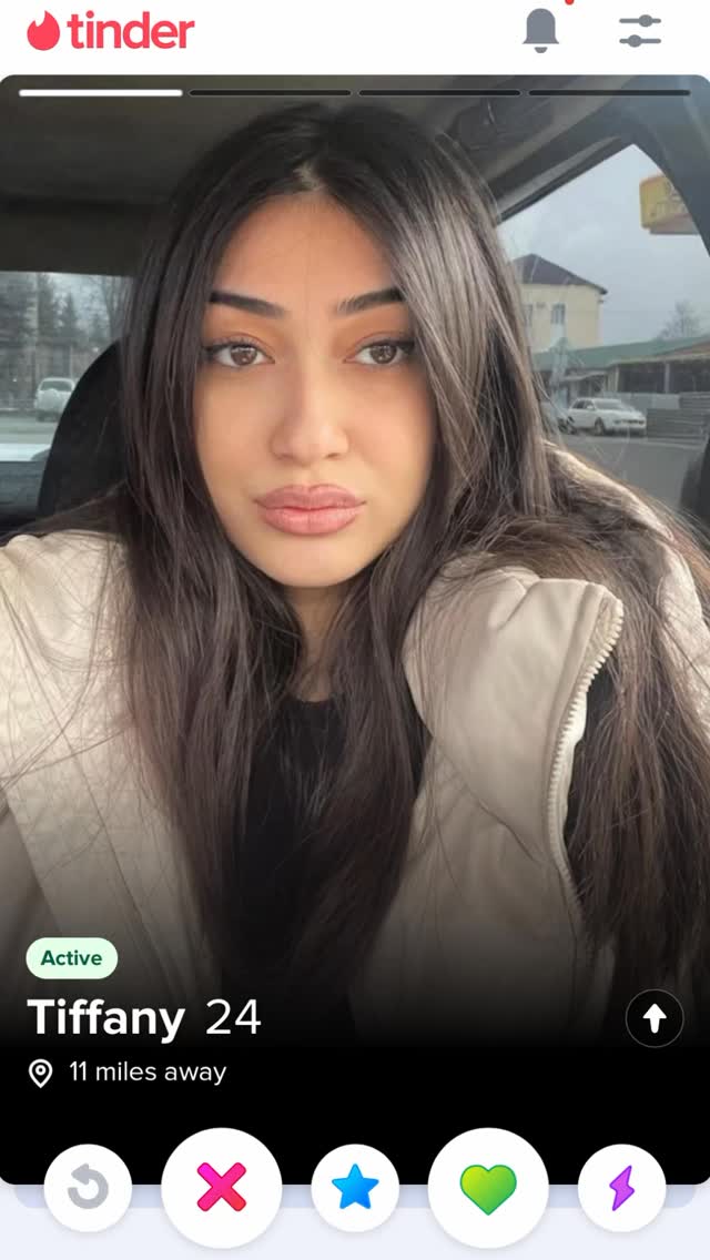 A screenshot from the dating app Tinder. The main focus is on a woman's face, with her hair and shoulders visible. The app interface shows her profile; the background shows a car and some blurred background elements.