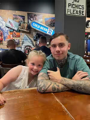 A photo taken at a restaurant shows a young man with extensive tattoos on his arms and neck seated at a table with a girl who has blonde hair. They are smiling and looking at the camera. The background shows a wall with a mural, posters, and a sign that says "Cold Beer."