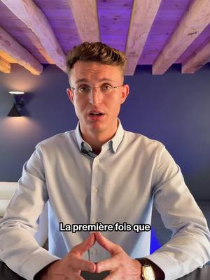 A man in a light blue button-down shirt with glasses is centered, looking directly at the camera. He's positioned against a gradient blue wall, with exposed wooden beams overhead and a modern black wall sconce to the left. Below him is text overlayed, saying 'La première fois que'.