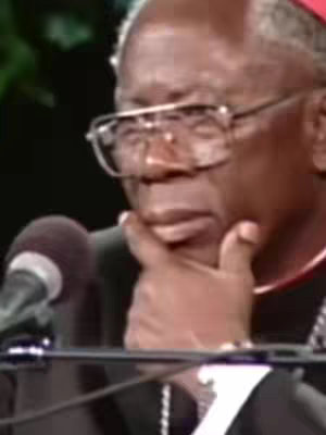 A close-up shot of an older Black man in glasses, holding his hand up to his chin as if pondering. He wears a dark suit with a partially visible white collared shirt, and a red collar can be seen. A microphone stand is partially visible to the left.