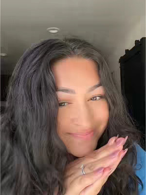A selfie-style image of a woman with long, dark hair. She is smiling slightly, with her hands clasped near her face, showing off a ring. The background is blurred, providing a soft, natural lighting.