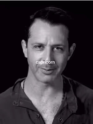 A black and white headshot shows a man with dark hair and a serious expression against a dark background. The word 'capricorn' is overlaid on his forehead. He is wearing dark clothing and a subtle chain necklace.