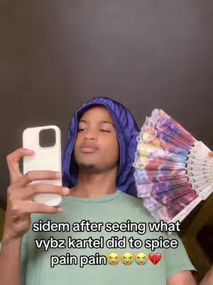 A close-up shot of a young Black man holding a phone and a decorative fan. He is wearing a blue head covering and a green t-shirt. There is text overlayed on the bottom. The background is dark.