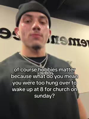 A selfie-style image featuring a young man looking up towards the camera against a background with large, white letters. He's wearing a black beanie, a black shirt, and a silver necklace with a cross pendant. Text overlayed on the image asks a provocative question about not being able to wake up for church.