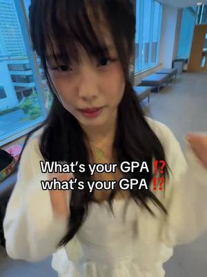 A young woman with long dark hair is centered in the frame. She wears a white top, and text overlays appear in the same font, asking, "What's your GPA ?!". The background is a blurred interior with a window showing city buildings, and a side bench.