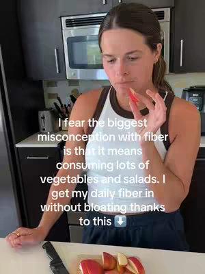A woman in a white tank top with a brown trim is looking down at a piece of red apple held in her right hand. She stands in a kitchen with dark cabinets and a microwave in the background. Text overlays the image.