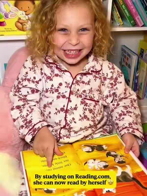 A young girl with curly blonde hair smiles widely while sitting in front of a bookshelf. She is wearing a floral pajama top and pointing at an open book with colorful illustrations. The text overlay has a yellow background and black font.
