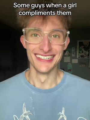 A young man with fair skin and short, light brown hair is centered in the frame. He smiles broadly, showing his teeth, and wears transparent-framed square glasses. He is wearing a light blue t-shirt with a bird graphic. The background is dark and blurred.