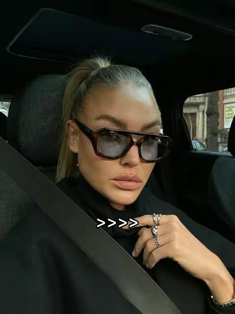 A woman wearing sunglasses, a black top, and rings sits in a car. The image is well-lit with a focus on the woman's face and upper body as she looks towards the camera.