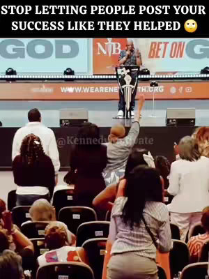 A wide shot from the audience perspective during a church service. The central figure is a preacher at a podium with a high-raised arm. In the foreground, people are seated, some with their hands raised, and there's a prominent sign behind the preacher.