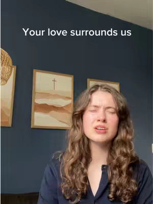 A woman with long, wavy brown hair and closed eyes is the central focus of the image. She appears to be singing or expressing emotion, with her mouth slightly open. Above her, the text "Your love surrounds us" is displayed. Behind the woman, the background consists of a dark blue wall, accented by two framed artworks with a cross in them. The image is well-lit.