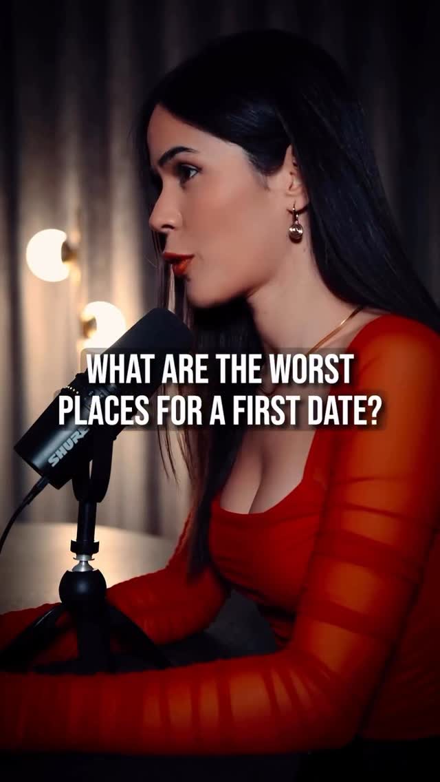 A woman with long black hair and red lipstick is speaking into a microphone. The image has warm lighting and a blurred background.