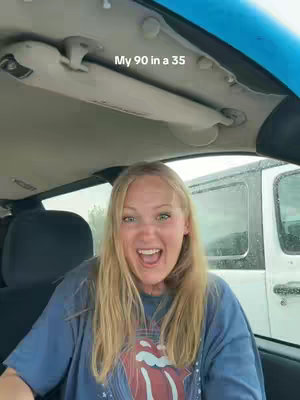 A woman with long blonde hair is sitting in a car, looking at the camera with a wide, excited expression. She is wearing a blue t-shirt with a graphic on it. The interior of the car is visible, with the ceiling and part of the windshield in the background.