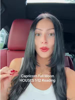 A woman with long, dark hair and red lipstick is seated in a car, looking directly at the camera. She is wearing a white top and a necklace. Text overlays read 'Capricorn Full Moon HOUSES 1-12 Reading.' The red car seat serves as the background.