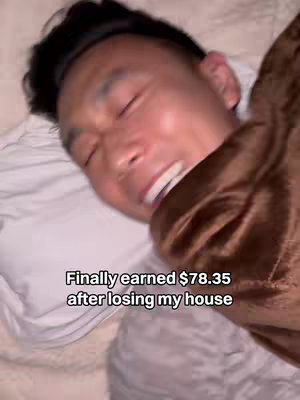 The image shows a close up of a man lying in bed. He is covered in a brown blanket and is smiling with his eyes closed. Below the face a text overlay says: Finally earned $78.35 after losing my house.