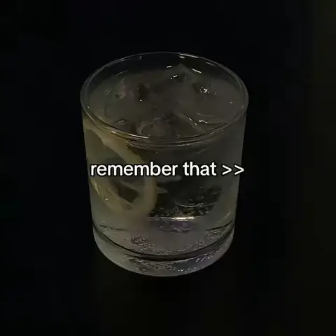 A clear glass containing a drink, likely a cocktail, with ice and a lemon slice, set against a stark black background. The image is darkly lit, emphasizing the contrast of the bright liquid against the shadows.