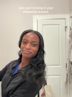 A young Black woman with long black hair is angled slightly to the side, smiling softly. She's wearing a black jacket over a blue shirt and holding a tote bag. The background is a neutral-colored wall and a white door.