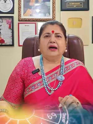 The video cover image features a woman, likely the creator, seated and speaking directly to the camera. She is in an indoor setting, possibly an office or studio, as indicated by the framed pictures on the wall behind her. The color scheme is warm, featuring a red sari which contrasts with the skin tone of the woman. Below her is a subtle overlay of zodiac-related graphics.