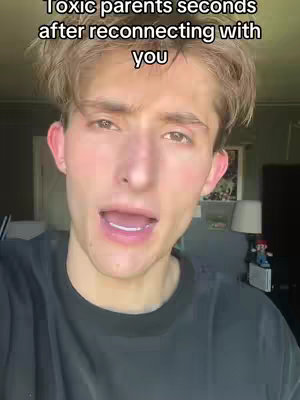 A young man with light brown hair and a black t-shirt is filming himself, looking directly at the camera with a slightly open mouth. Behind him are a picture on the wall, a lamp and light coming through.