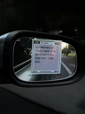 The image shows a car's side mirror reflecting a smartphone screen. The screen displays a text message in a bubble, within the reflection the road and sky are visible. The overall setting suggests a driving scene.
