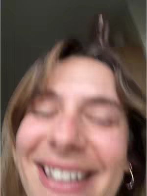 A blurry close-up shot of a woman smiling with her eyes closed. The focus is on her face, with her hair partially obscuring the top of the frame. The background is out of focus, suggesting an indoor setting with soft, natural lighting.