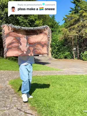 A person stands in front of a tapestry or large fabric item, obscuring their upper body. They are wearing light blue jeans and white sneakers. The background includes green grass, trees, and a bright blue sky.