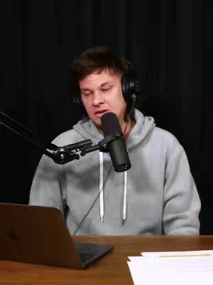 A man with short, dark hair wearing a gray hoodie and headphones sits in front of a microphone, looking down with a somewhat concerned expression. A laptop and papers are on the desk in front of him and the background is dark.