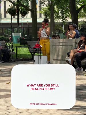 An outdoor scene with a white card overlaid in the foreground. The card features a question in bold text, with a blurred figure standing behind it and other people sitting on benches in the background. The color palette is dominated by natural greens for the trees and grass.