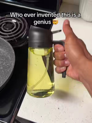 A person holding a clear bottle filled with yellow liquid, likely oil for cooking. They are giving a thumbs-up. The scene is set in a kitchen, with a stove and countertop visible in the background.