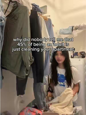 A woman is standing in what appears to be a cluttered closet or storage space, holding up a stack of folded clothes. The space is filled with hanging clothes and other items, suggesting a laundry or cleaning context. The background includes a washing machine and other household storage items.