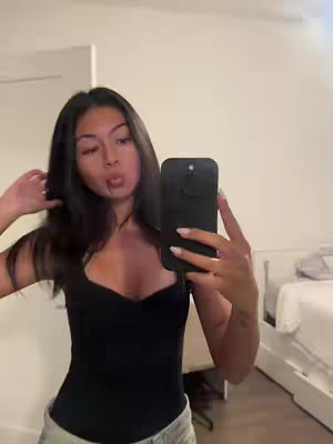 A young woman of Asian descent is taking a selfie in a well-lit room. She has long, dark hair that is partially covering her face. She's wearing a black top and holding a black phone with her left hand. Her lips are puckered slightly, and she appears to be making a playful face. Light-colored walls and part of a bed with a white bedspread are visible in the background. The composition is natural and casual, with the woman looking directly at the camera.