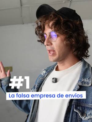 A man in glasses and a black cap is gesturing with his right hand while speaking. Text overlays read '#1' and 'La falsa empresa de envíos' in a white bar. The man wears a white t-shirt and a denim jacket with a microphone attached. The backdrop is a plain white wall.