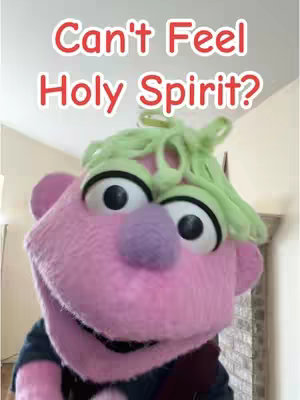 A close-up of a pink puppet with a large face, big black eyes, and green hair. The puppet is positioned in front of a neutral indoor background, with text overlayed at the top.