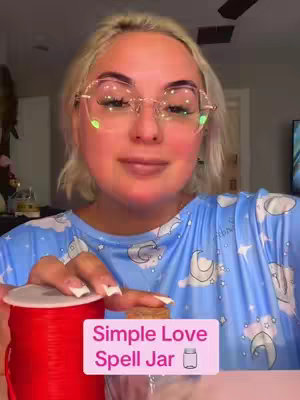 A woman with blonde hair and glasses is holding a small jar, a roll of red string, and a cork. The image also features text that reads 'Simple Love Spell Jar'.