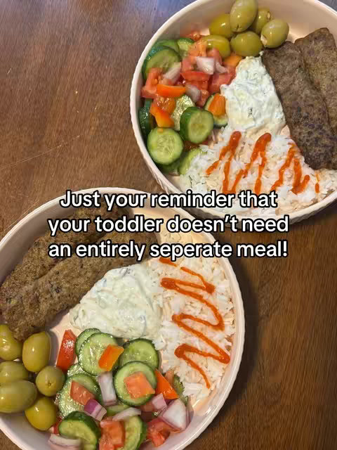 A top-down shot showcases two white oval-shaped ceramic bowls filled with Mediterranean food, placed on a wooden table. Each bowl contains a mix of kebab, rice, cucumber and tomato salad, tzatziki sauce, and olives. The bowls are positioned towards the viewer, emphasizing the food. The text overlay, 'Just your reminder that your toddler doesn't need an entirely seperate meal!', is in the center of the image.