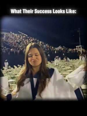 A young woman, wearing a white choir robe with dark blue accents, stands in front of a large crowd, smiling slightly with her eyes looking down. The setting appears to be a night-time outdoor event, possibly a graduation ceremony, with rows of chairs and other people in the background. The color scheme consists primarily of dark tones due to the lighting conditions.