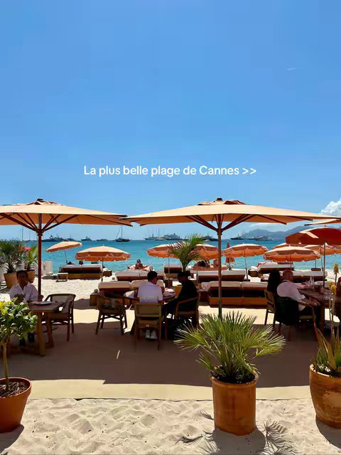 A sunny beach scene with a clear blue sky. Several large, open umbrellas provide shade for tables and seating areas filled with people enjoying the day. The sea and some boats are visible in the background. The foreground features a sandy area with potted plants.