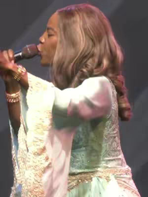 A woman with long blonde hair wearing a light turquoise dress and holding a microphone, appearing to be singing on stage.