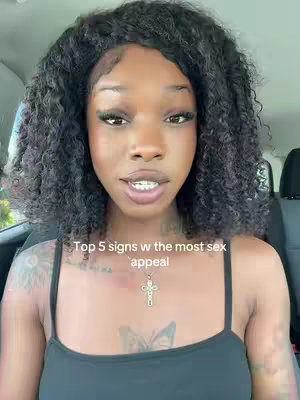 A selfie style shot of a woman in a car. She has dark, curly hair and is looking directly at the camera. She wears a black top and a cross pendant necklace. Text overlay in the bottom center reads, "Top 5 signs w the most sex appeal."