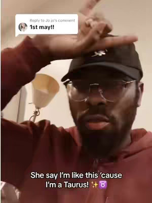 A selfie-style video featuring a Black man wearing a black cap and glasses smiling. He is wearing a maroon shirt, with his arm raised and partially obscuring his face. A comment reply is displayed above his head.
