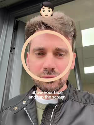 A selfie-style shot of a man's face, centered in the frame. A graphic of a circle is overlaid on his face. A small cartoon avatar sits atop his head. The background appears to be a semi-industrial setting with dark gray walls and a window or door in the back.