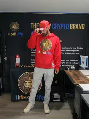 A man in a red hoodie and a red baseball cap stands facing forward, holding his hand up to adjust the angle of his hat. The background is a black backdrop with white text and a large gold IHodlLife logo. He is also wearing light grey pants and white sneakers. In the background there's a table with the IHodlLife logo. The scene is lit by a fluorescent light.