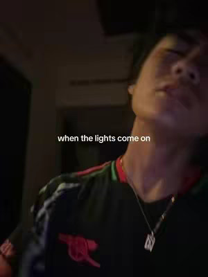 A selfie-style shot featuring a person from the chest up, seen in dim lighting. The person is wearing a dark top with sleeves, appearing to be a jersey with red and green accents and an emblem. The person's face is partially visible, with the focus on the upper body and the face tilting up. Text overlay in white font displays 'when the lights come on'.