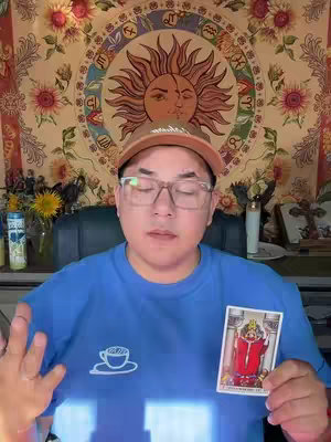 A man with glasses and a hat holds a tarot card against a background adorned with tapestry and sunflowers. He is wearing a blue shirt with a coffee cup logo. Overall, the scene seems to be set up for a reading or prediction.