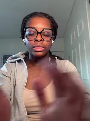 A young Black woman with glasses and braided hair, wearing a gray hoodie and a beige top. Her hands are in front of the camera. The background shows a room.